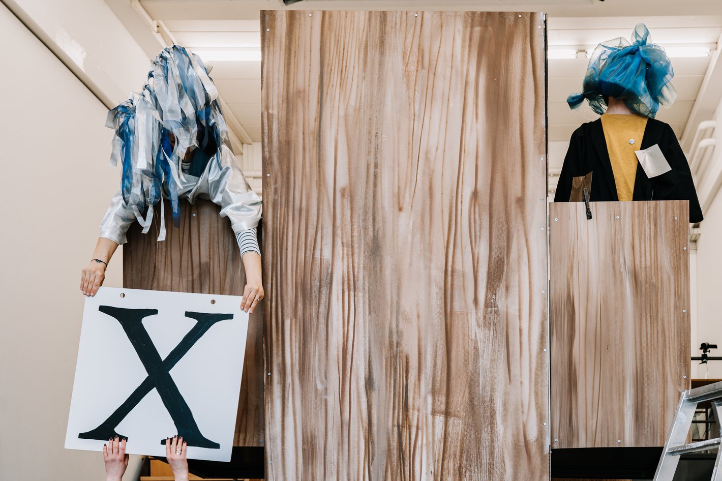 Zwei mit Stofffetzen vermummte Personen stehen auf und hinter einem Holzpodest. Die linke Person nimmt ein Schild mit einem großen "X" an, während die andere steif steht.