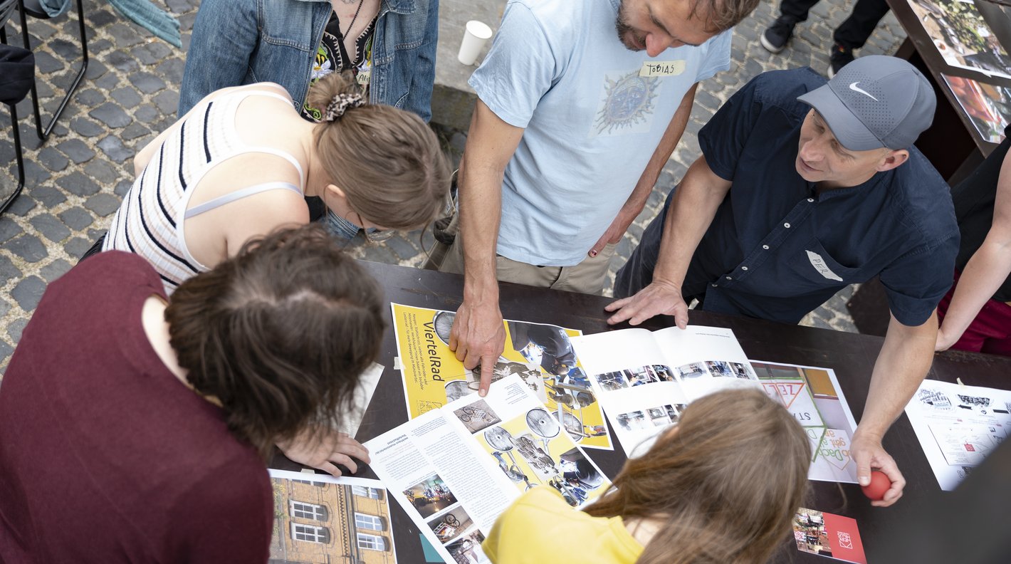 Personen die während des Projekt "Trans Urban X" um einen Tisch herum stehen und miteinander reden. Auf dem Tisch sind Broschüren und Fotos ausgebreitet.