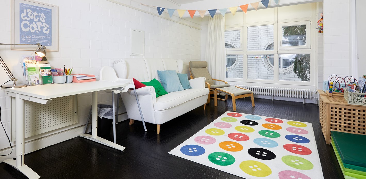 Foto des Familienzimmers mit Arbeitsplatz, Sofa, Spielteppich, Spielzeug, Büchern, Stillecke und Wickelplatz