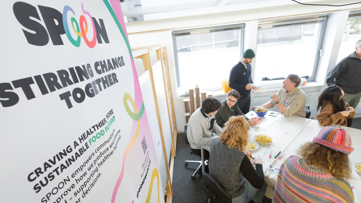 Studierende mit dem Team von SPOON arbeiten an einem Tisch zusammen. Es wird auf die Tischdecke gezeichnet und nachhaltig gekocht.