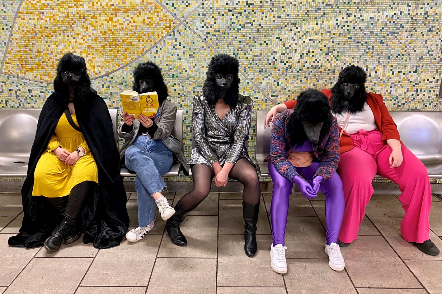 Fünf Personen mit Hundemasken und bunt gekleidet sitzen auf einer Bank in einer U-Bahnstation.